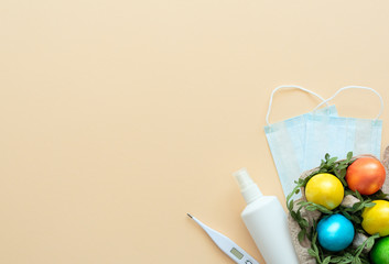 Decorated Easter eggs lie in the carton box on yellow background with medical face mask, thermometer and sanitizer. Happy Easter during quarantine, stay home, social distance, COVID-19 concepts.