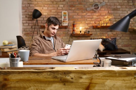 Casual man using mobilephone, sitting at desk, having laptop, texting.