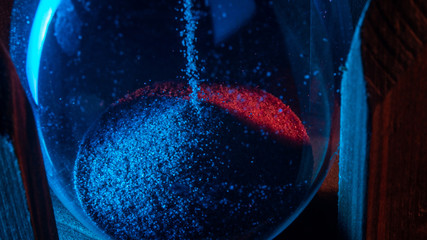 a trickle of sand falls into the lower vessel of the hourglass. close up. blue-red color. abstract view. black background