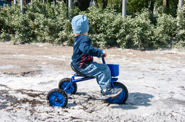 A blond-haired boy in blue rides a small tricycle in a puddle completely covered with the old...