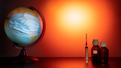 A medical mask is worn on an earth globe. World quarantine, coronavirus pandemic. Orange gradient background. Vials of medicine and a syringe