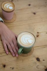 Hand with green cup with coffee on wooden desk. Top view in cafe.