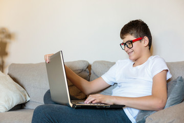 Cute young boy in glasses and white t shirt sitting on the couch in the living room with laptop and study. Homeschooling, self education, distance learning by kids.	