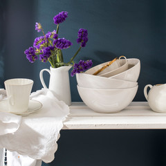 A bouquet of limonium and a set of white dishes: porcelain milk jug, coffee cup, bowls on a white rack on a dark blue background