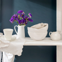 A bouquet of limonium and a set of white dishes: porcelain milk jug, coffee cup, bowls on a white rack on a dark blue background