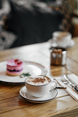 A white cup of coffee. A delicious dessert of soufflé and berries on a round biscuit layer lies in a white plate sprinkled with powdered sugar. A photo with a blurred background.