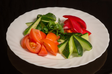 Sliced vegetables-cucumbers, tomatoes, red pepper and parsley on a white plate. against a black reflective surface