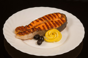 a piece of grilled salmon with lemon and olives on white plate. On the background of black reflective surface