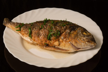 grilled whole fish with seasonings on a white plate. On the background of a reflective surface