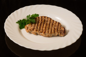 a piece of grilled pork with a sprig of parsley on a white plate. On a black reflective table