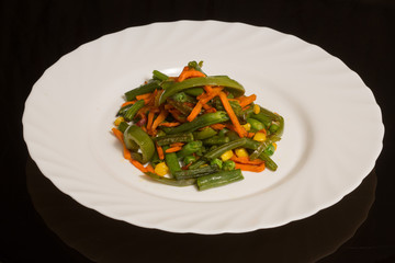 A dish of passaged vegetables on a white plate. string beans, green peas, corn, peppers, carrots. against a black reflective surface
