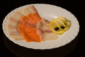 sliced fish delicacies on a white plate. oil fish, trout with lemon. against a black reflective surface
