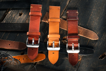 Handmade brown tone watch straps with steel buckle laying on wooden rustic surface next to leather thread. Different leather colour tones.