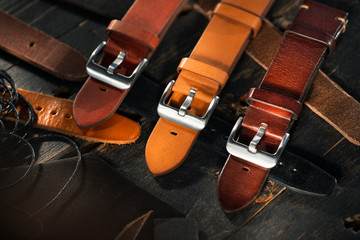 Fototapeta na wymiar Close up different colour watch straps with steel buckles laying next to the leather cuttings and leather thread. Dark wooden surface.