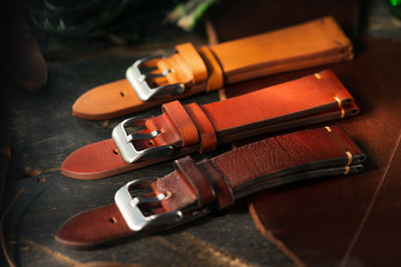 Handmade brown tone watch straps with steel buckle laying on wooden rustic surface next to leather cuttings and leather thread. Different leather colour tones. - Powered by Adobe