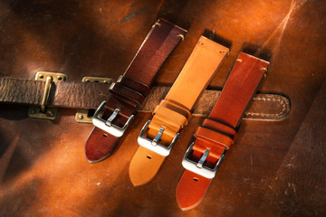 Handmade brown tone watch straps with steel buckle laying on wooden rustic surface next to leather cuttings and leather thread. Different leather colour tones.
