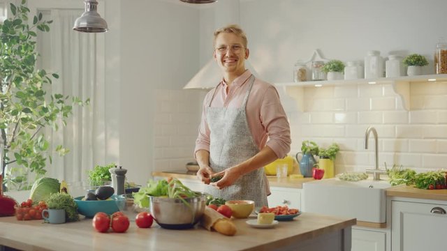 Handsome Man in Pink Shirt and Apron is Making a Healthy Organic Salad Meal in a Modern Sunny Kitchen. Hipster Man in Glasses Smiles at the Camera. Natural Clean Diet and Healthy Way of Life Concept.