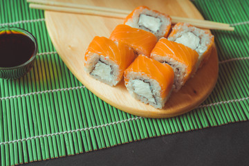 Delicious rolls on wooden board with soy sauce on decorative bamboo napkin. Close up of tasty sushi rolls with chopsticks on cutting board on black background.