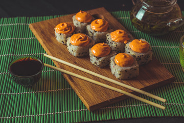 Delicious rolls on wooden board with chopsticks and green tea in teapot on decorative bamboo napkin. Close up of tasty sushi rolls on cutting board on black background.