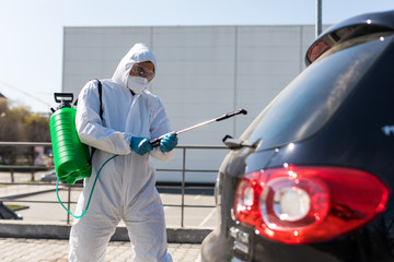 Coronavirus Pandemic. Disinfector in a protective suit and mask sprays disinfectants of car outdoors