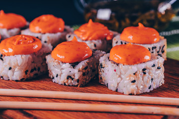 Delicious rolls with spicy sauce on wooden board on decorative bamboo napkin. Close up of tasty sushi rolls with chopsticks on cutting board.