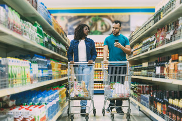 Peaceful young couple making purchases in grocery store. Bearded man in eyeglasses holding smartphone. Young woman with dreadlocks looking at boyfriend. Shopping concept