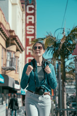 young girl photographer with backpack and camera relax walking on street in castro area. smiling woman in sunglasses sightseeing in rainbow district in san francisco california on sunny blue sky.