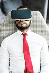Serious businessman in virtual reality headset. High angle view of concentrated young African American businessman sitting in chair and using VR headset in office. Technology concept