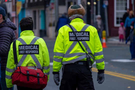 Two medical first responders walking in a street with people in the background. The officers are wearing a bright yellow reflective coat with grey stripes and a red first aid bag. 