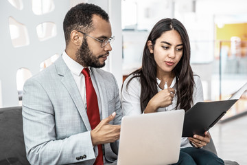 Obraz premium Coworkers with folder and laptop discussing work. Professional multiethnic businessman and businesswoman working with laptop computer and documents in office. Cooperation concept