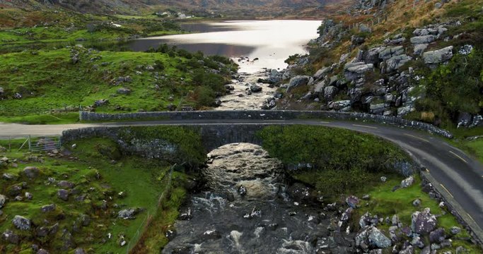 Stunning aerial fly-over Kerry Road in Ireland. Stone bridge crossing the river. Surrounded by hills with rocks.