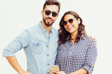Obraz premium Positive couple of tourists wearing sunglasses, using smartphone, smiling at camera. Young woman in casual and man in glasses in glasses posing isolated over white background. Vacation concept