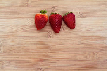 Three strawberries arranged in a row on a bamboo cutting board; strawberries as a top boarder on a background of bamboo