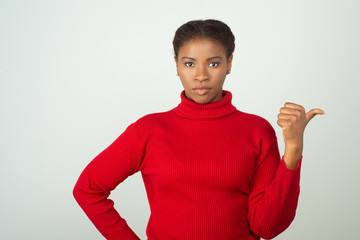 Serious black woman wearing red, pointing thumb at copy space. Beautiful young African American lady standing isolated over white background. Advertising concept