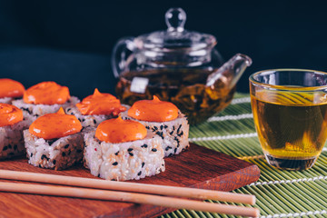 Delicious rolls with spicy sauce on wooden board with green tea in teapot and cup on decorative bamboo napkin. Close up of tasty sushi rolls with chopsticks on board on black background.