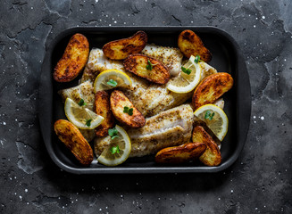 Baked fish fillet and potatoes on a baking sheet on a dark background, top view. Mediterranean style lunch