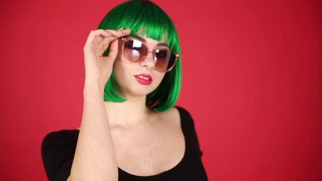 Beautiful girl in a green wig and pink glasses grimaces, fools around and expresses different emotions. Video