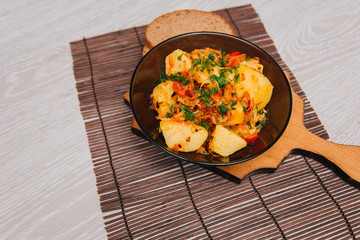 Stewed potatoes with cabbage in a dark plate on a brown board, on a light wooden table. Rustic cuisine.