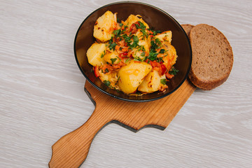 Stewed potatoes with cabbage in a dark plate on a brown board, on a light wooden table. Rustic cuisine.