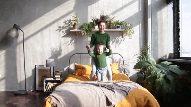 Mom and daughter dressed equally in green sweaters and jeans are jumping on the bed. Slow motion video