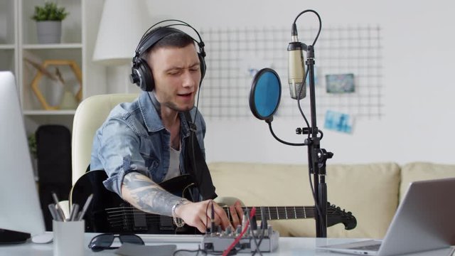 Young tattooed man in headphones playing the guitar, using console and singing into microphone while recording song at home