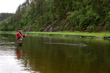 Side view portrait of brutal aged fisherman standing hip-deep in narrow forest river at the moment of catching fish by fishing tackle. Active hobby