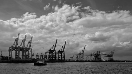 Obraz premium Panoramic view of the Port of Hamburg with cranes on the background along the river and a boat on water, under a cloudy sky, in black and white.