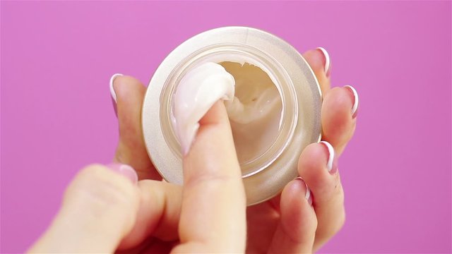 woman hand takes moisturizer cream from glass jar with her finger extreme close up pink background