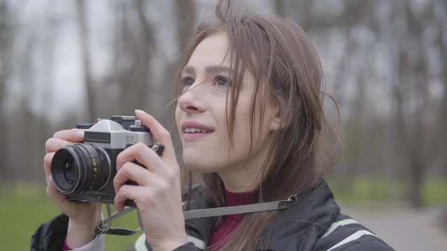Close-up portrait of positive female photographer with camera. Young excited woman taking photos of nature in park. Brunette brown-eyed girl enjoying hobby outdoors. Leisure, lifestyle, creativity.