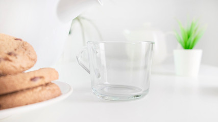 Pouring milk into glass on kitchen table with cookies. Close up.