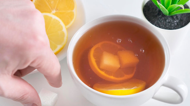 Add sugar to whute cup of tea on kitchen table. Close up.