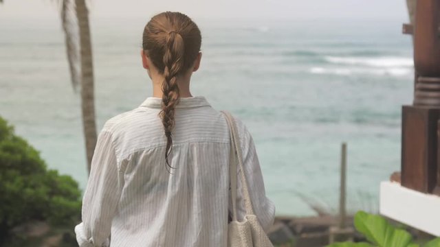 young woman walks near rest hotel against tropical plants and storming ocean backside view slow motion