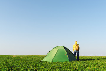 Tourist in yellow jacket near his tent on summer field. Clear blue sky. Travel and adventure concept