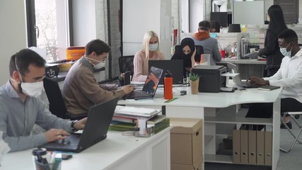 International office, working and communicate in an open space, office workers and managers in medical masks working at computers, protection from the virus, working during the pandemic. - Powered by Adobe